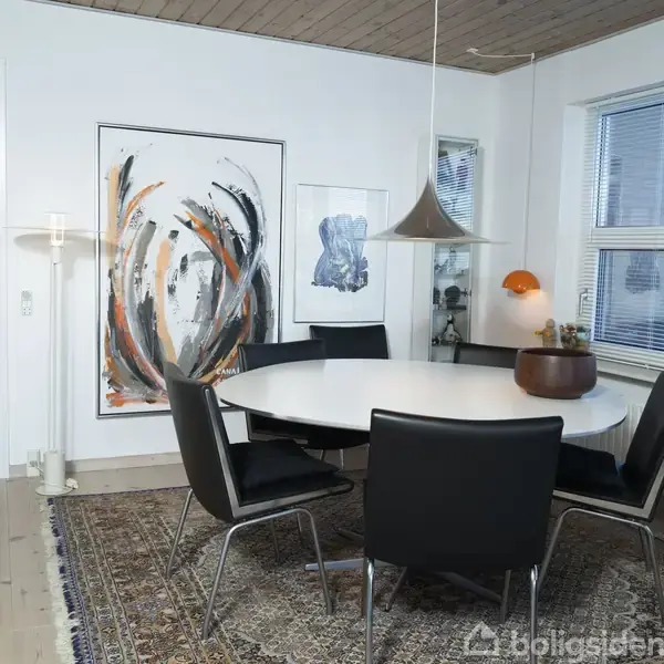 Et rundt, hvidt spisebord med seks sorte stole står på et orientalsk tæppe. En moderne lampe hænger over bordet, omgivet af kunstmalerier. Vinduer fylder rummet med lys.