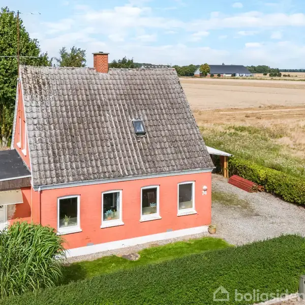 Et rødt hus med gråt tegltag står omgivet af en velplejet have med græs og buske. Det ligger ved siden af åbne marker under en skyet himmel.