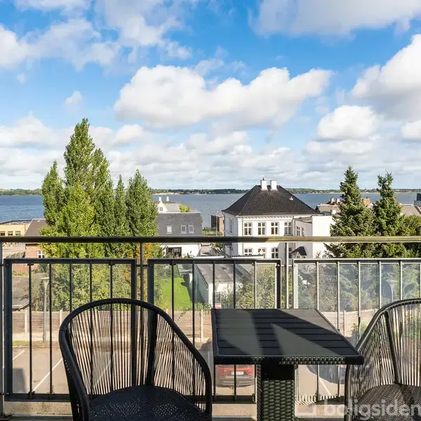 To stole ved et bord står på en balkon med udsigt over vand og træer. I baggrunden er der bygninger og en blå himmel med skyer.