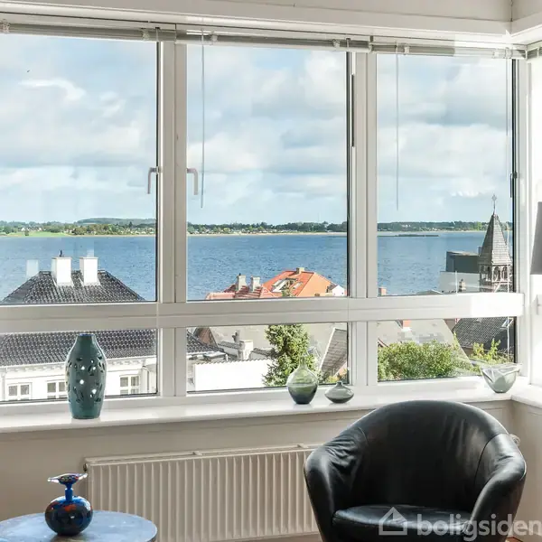 Et stort vindue med udsigt over en by og havet. Foran vinduet står en sort lænestol, et bord med en blå vase og en lampe.