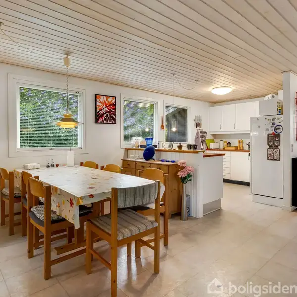 En køkkenø med orange bordplade står centralt; omgivet af lyse vægge og vinduer. I baggrunden ses en stue med sofagruppe og store vinduer, der lukker lys ind.