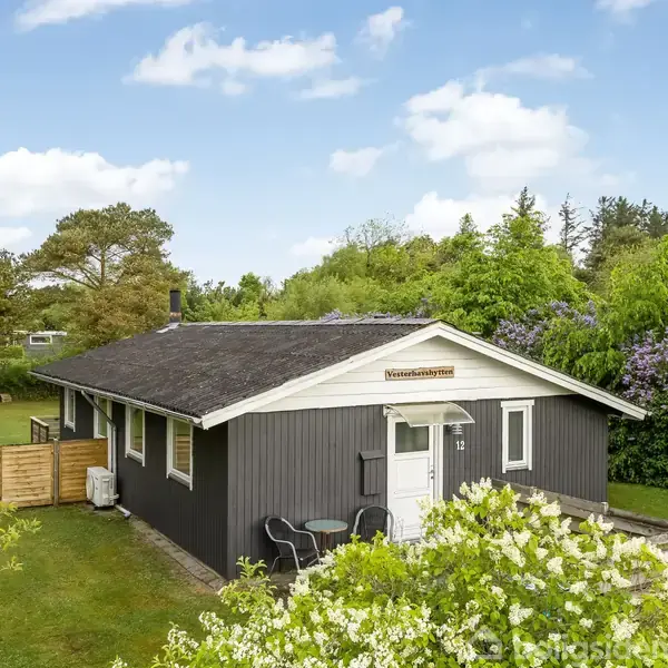Mørkt træsommerhus med skilt "Hammerstien 19". Det står i en frodig have med diverse buske og træer. Foran huset er der et lille rundt bord med to stole.