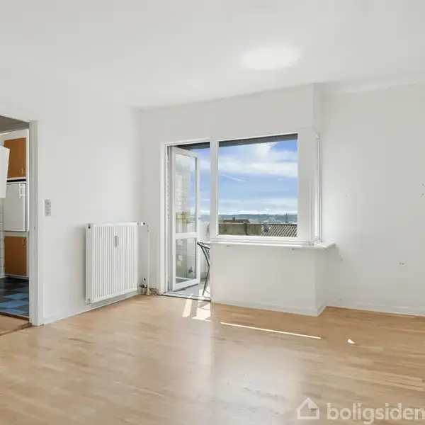 Stue med parketgulv og hvidmalede vægge; glasdør åbner mod altan; radiator placeret ved siden af døren; døråbning til køkken med flisegulv. Blå himmel ses udenfor.
