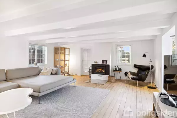 En moderne stue med en stor grå sofa står midt i rummet. En pejs er tændt, og lyse trægulve strækker sig ind i rummet. Møblerne er minimalistiske, og dagslyset stråler ind gennem vinduerne.