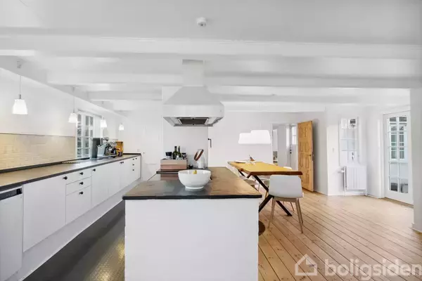 Et moderne køkken med en central køkkenø, sunde skåle og en hætte i centrum, omgivet af hvide overflader og træplankegulv. I baggrunden ses et spisebord og stole.