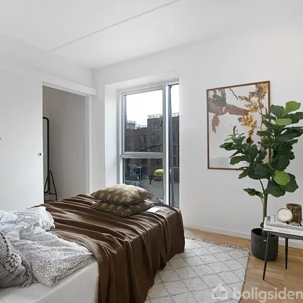 Soveværelse med en seng dækket af puder og brun sengetæppe. En stor grøn plante står ved siden af et lille bord. Et åbent vindue i baggrunden lader lys ind.