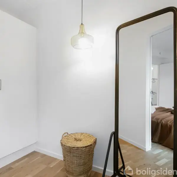 En stor gulvspejl står oprejst i et lyst rum ved siden af en lukket kurv. Til venstre ses et skab. En lampe hænger fra loftet.