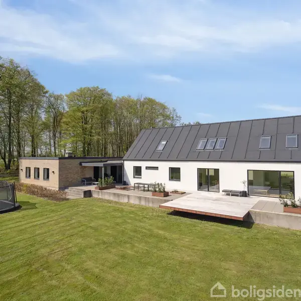Et moderne hus med hvide og træfarvede facader står på en stor, grøn græsplæne. En trampolin står i haven, og i baggrunden ses træer mod en blå himmel.