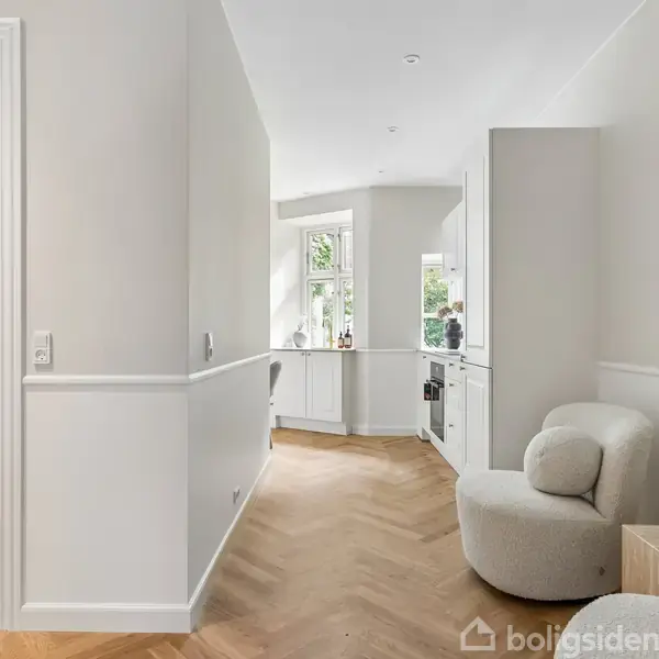 Et lyst, elegant køkken med træparketgulv strækker sig ned ad en gang. Til højre står en rund lænestol ved et lille bord med en vase og stearinlys.