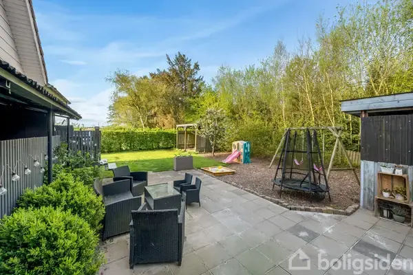 Terrasse med sorte havemøbler står stille ved siden af et gyngestativ og sandkasse. Bag ved ses en grøn græsplæne omgivet af træer og buske under en klar blå himmel.