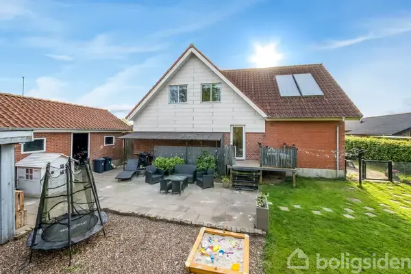 Hus bagfra med rødt tag og hvid facade med en terrasse med havemøbler. Til venstre, en trampolin; foran, en sandkasse. Græsplæne omgiver området under en solrig himmel.