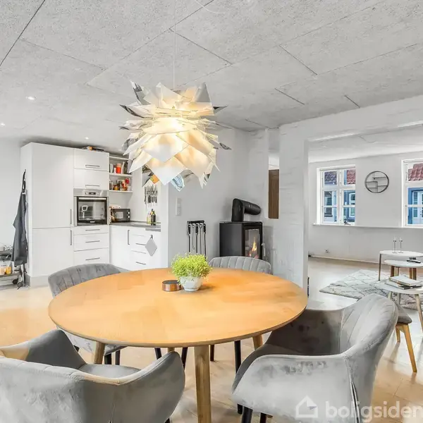 Et rundt træbord omgivet af grå stole står i en moderne, lys stue-køkken-alrum med parketgulv og en skulpturel lampe. I baggrunden ses et hvidt køkken og store vinduer.
