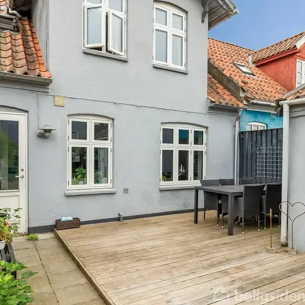 Grå bygning med rødt tag omkranser en træterrasse med et mørkt spisebord og stole. Nabohuse med farverige facader ses i baggrunden. Atmosphäre er hyggelig og indbydende.