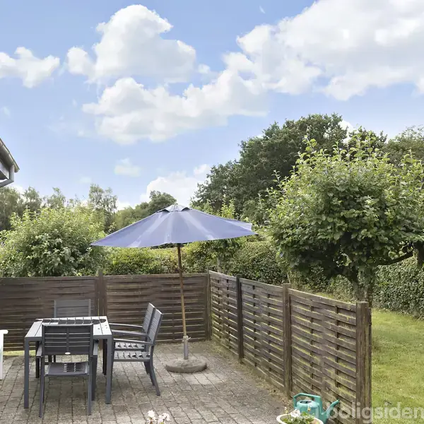 Terrassemøbler står under en blå parasol på en flisebelagt terrasse. Haven er omringet af grøn bevoksning. Vejret er solrigt med blå himmel og nogle skyer.