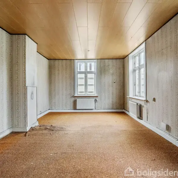 Et tomt rum med mønstret tapet viser slidte gulvtæpper. Tre vinduer giver naturligt lys til rummet, mens en radiator er placeret under hvert vindue. En lille kasse eller skab findes i hjørnet.