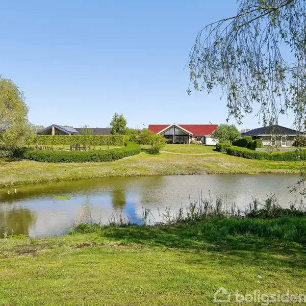 Huse med røde tage står stille ved en dam omgivet af grønne træer og græs. Bunken af træer og spejlende vand skaber en fredfyldt scene i en landsby.