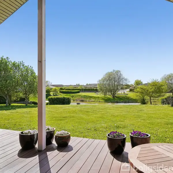 Træterrasse med blomsterkrukker står stille, omkranset af en velplejet græsplæne og flere træer, under en klar blå himmel i en fredelig have.