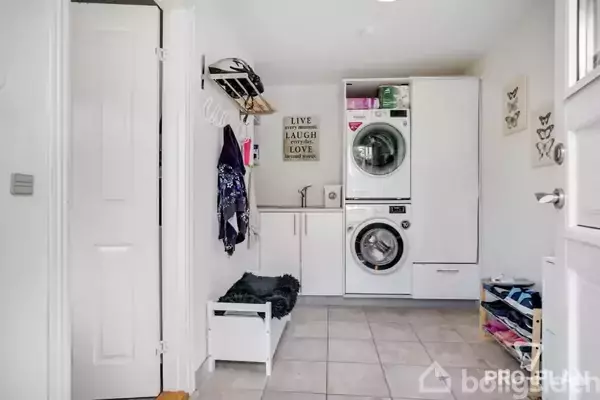 Vaskemaskine og tørretumbler stablet i et bryggers med skabe og flisegulv. En bænk står ved siden af en knagerække. Tekst på væggen: "LIVE LAUGH LOVE."