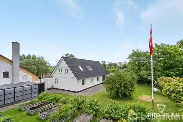 Et hvidt hus med sort tag står i en frodig have med bede og træer. Til højre vajer et dansk flag på en flagstang.