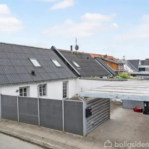 Et hvidt hus med skråt tag står ved en stille vej. Carporten er delvist åben, omgivet af stakit og andre huse i baggrunden. Skiltet viser "30 P".