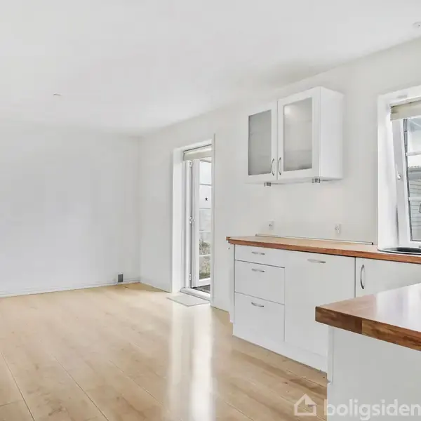 Køkken med hvide skabe og træbordplade; vinduer til højre giver lys; dør fører ud til gårdhave. Rummet har lyse trægulve og hvide vægge.