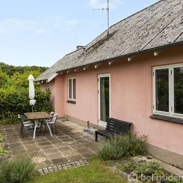 Et lyserødt hus har en terrasse, hvor et bord med stole står. Parasol skaber skygge. Omkranset af en grøn have med buske.