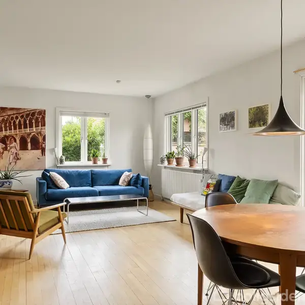 En stue med en blå sofa står foran to vinduer med udsigt til en grøn have. Et rundt spisebord med stole er ved siden af en reol fyldt med bøger.