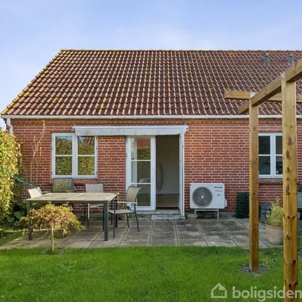 Rødt murstenshus med skråt tag og flisebelagt terrasse. Terrassebord og stole står ved siden af en hæk. En varmepumpe er monteret ved husets mur. Græsplæne og pergola omgiver området.