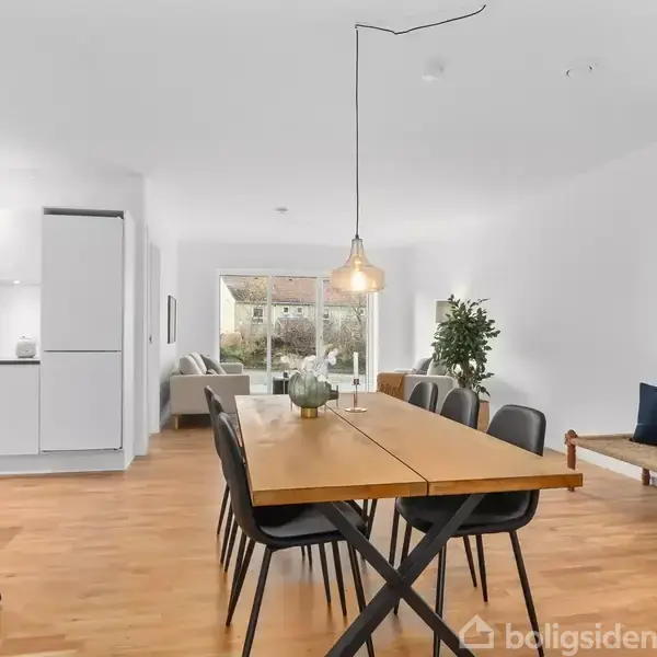 En spisestue med et langt træbord omgivet af sorte stole, står under en hængende lampe. I baggrunden ses et køkken og opholdsområde med et vindue og planter.