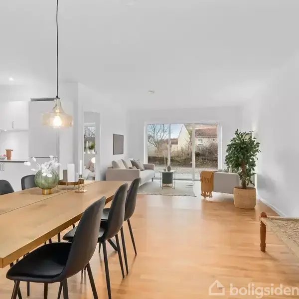Langt spisebord med seks stole står i en rummelig, lys stue. I baggrunden er et opholdsområde med sofa og store vinduer mod en have. Træmøbler og grønne planter skaber en varm atmosfære.