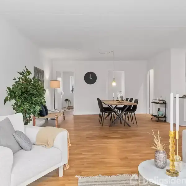 Stue med hvide sofaer i forgrunden, dekoreret med puder og tæpper, vender mod et spisebord med sorte stole. Lyst trægulv og en stor plante i hjørnet.