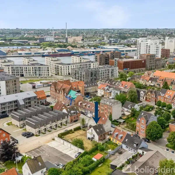 Bygninger står stille i en tæt bebygget by, omgivet af boligblokke og industribygninger ved en fjord. Det bagvedliggende landskab er grønt og kuperet.