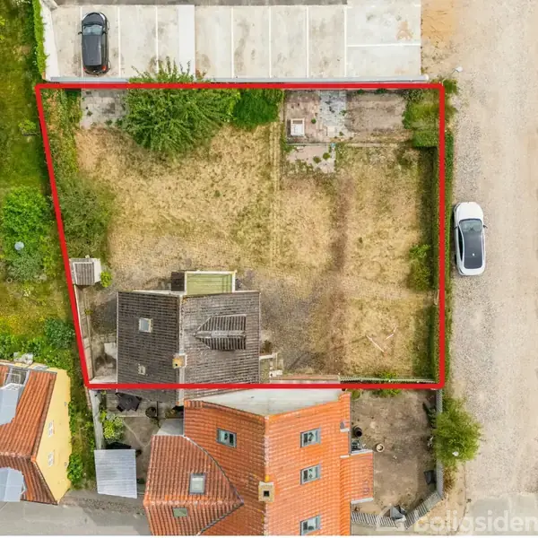 En grund med tørt græs er indrammet med en rød linje. En bygning står på grunden. Parkeringspladser og flere biler er i nærheden.