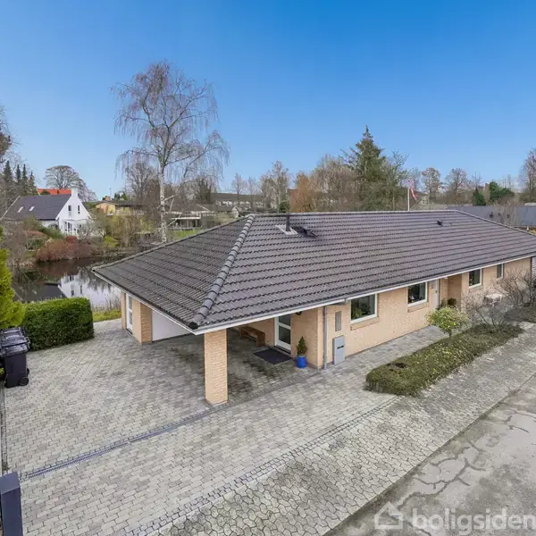 En etplans villa med gråt tegltag, omgivet af en flisebelagt indkørsel og havebede, står ved en rolig vej. I baggrunden ses en lille sø og flere træer.