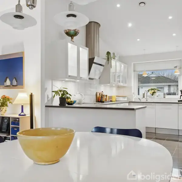 En skål står på et hvidt spisebord i et moderne køkkenmiljø. Køkkenet har hvide skabe, en emhætte, planter og forskellige dekorationer, såsom malerier og lamper. En stor vindue tilføjer lys.