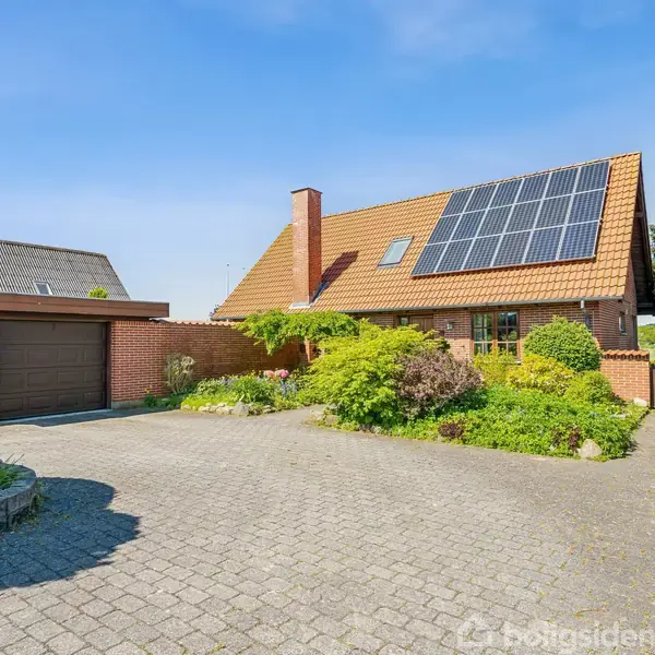 Et rødt murstenshus med solpaneler på taget står i en velplejet have. En flisebelagt indkørsel leder op til en garage. Scenen er under en klar blå himmel.