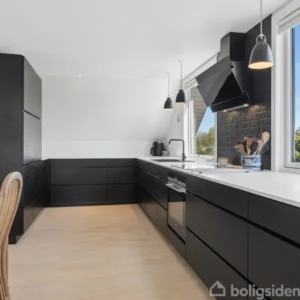 Sort, moderne køkken med hvide bordplader. Køkkenet har en emhætte over komfuret, store vinduer med udsigt til grønt område, og der hænger flere sorte lamper fra loftet.