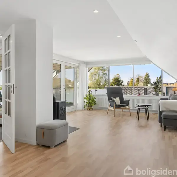 Stue med en lænestol og sofabord, indrettet i lyst, moderne design. Rummet har skråvægge, vinduer med udsigt til træer, og en glasterrassedør. Ingen tekst er synlig.