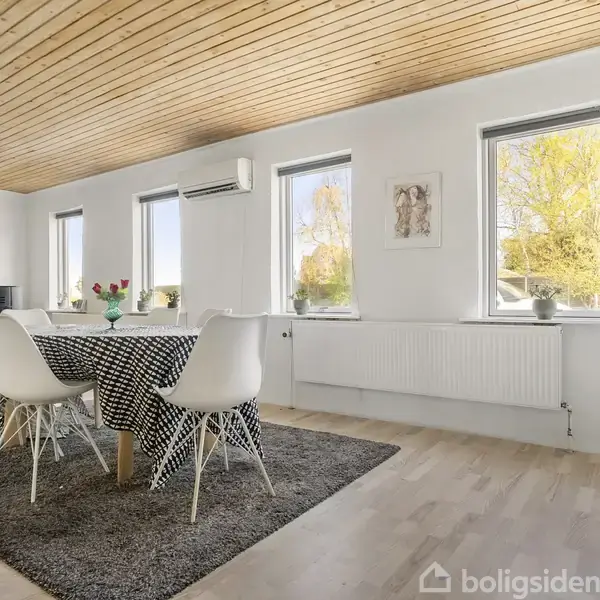 Spisebordet er omkranset af hvide stole, står på et gråt tæppe, med en sort-hvid mønstret dug. Bagved ses tre vinduer, en sofa, og træloft i moderne stue.
