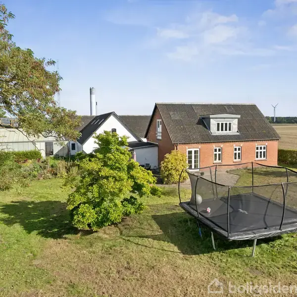 Hus med rødt murværk og sort tag står i en have. En trampolin står på græsplænen. I baggrunden er der marker under en blå himmel. Træer og buske omkranser ejendommen.