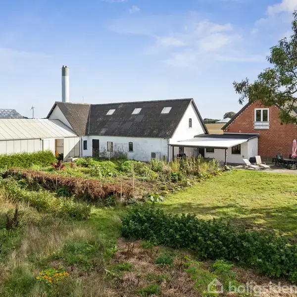 En hvid og rød murstensbygning står i en landlig have med et drivhus og en græsplæne. Træer omgiver området, og en terrasse med møbler ses til højre.
