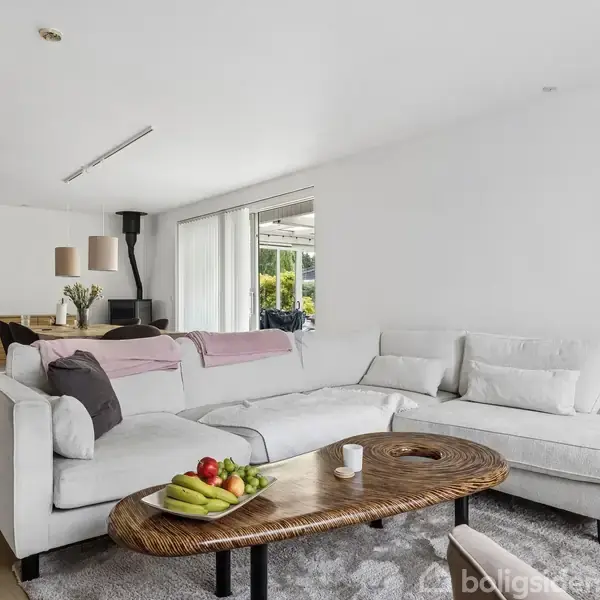 En stor, lys sofa står i en stue med et sofabord foran. Der ligger frugt på bordet. Store vinduer giver udsigt til en terrasse og have.