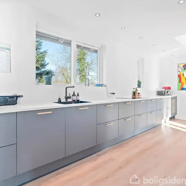 Et moderne køkken med grå skabe strækker sig langs en væg. Et stort vindue giver udsigt til træer, mens farverige kunstværker dekorerer væggen. Sollys strømmer ind.