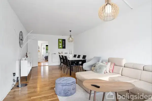 En beige hjørnesofa står i en moderne stue med trægulv. Et rundt sofabord og en lilla puf er placeret foran den. Et spisebord med sorte stole ses bag sofaen.