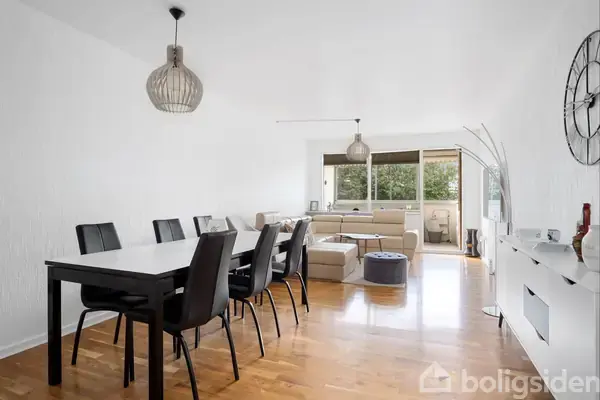 Spisebord med sorte stole står i en lys spisestue; to lamper hænger over bordet. I baggrunden er der en sofa ved store vinduer med udsigt til grønne træer.