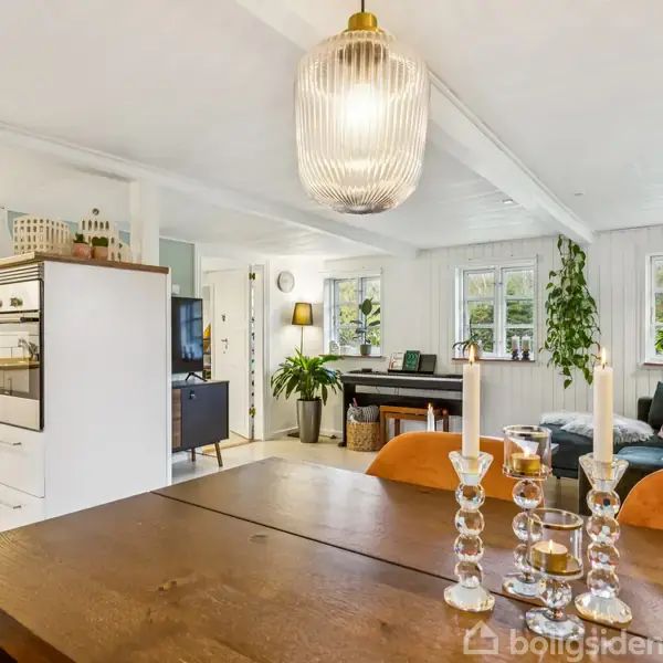 Lysestager med stearinlys står på et træbord i en stue med sofa, planter og klaver. Et åbent køkken ses i baggrunden med hvide skabe og hylder.