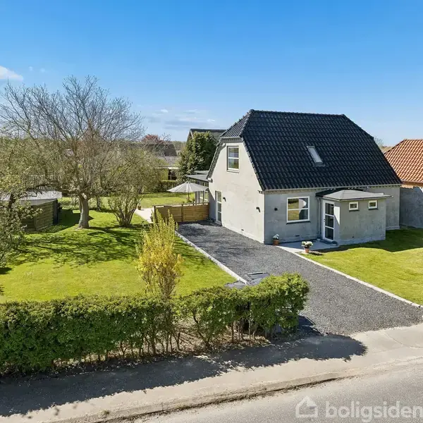 Et gråmalet hus med sort tag står på en velholdt græsplæne; omgivet af træer og buske, ses ved en vej. I baggrunden er der udsigt over vandet under en blå himmel.