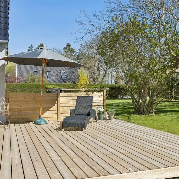 Terrasse med solseng under parasol. I hjørnet står et lille bord med stol. Haven omkring har grønne græsplæner og træer, huset ses til venstre.
