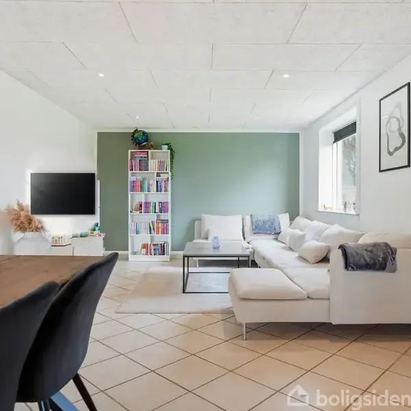 En lys stue med en hvid hjørnesofa mod en grøn væg. Et fladskærms-TV hænger ved siden af en reol fyldt med bøger. To stole står ved et træbord.
