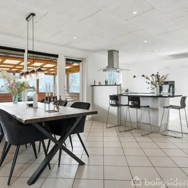 Spisebord med sorte stole står centralt i en moderne, lys spisestue. Ved siden af ses et åbent køkken med barstole og ø-station. Store vinduer giver udsigt til terrasse og have.